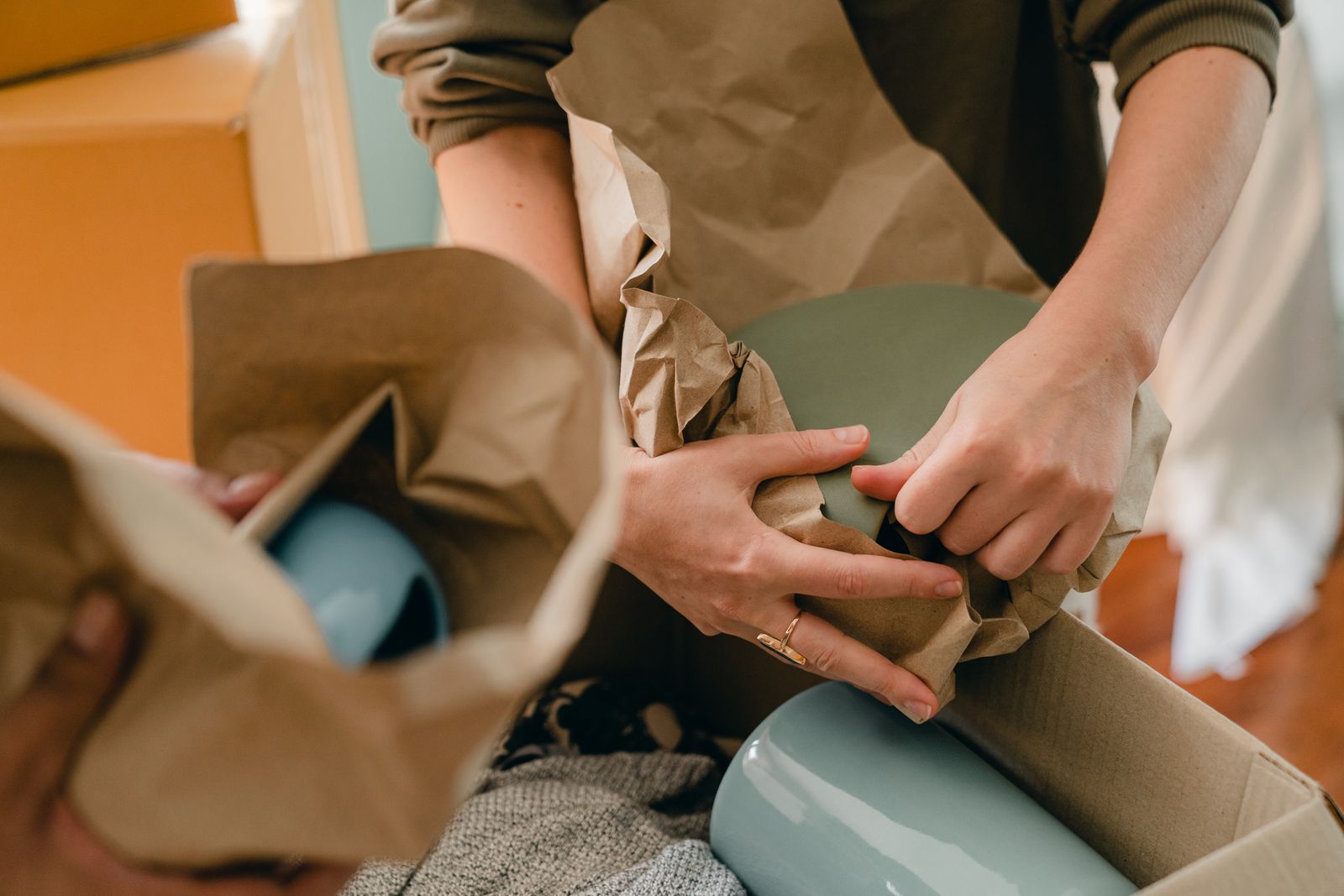 Hands protecting a ceramic plate with packing paper above a moving box
